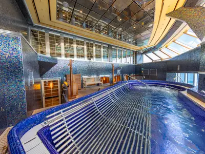 Ein luxuriöser Spa-Wellnessbereich auf der Carnival Luminosa. Geschwungener Pool mit blauen Mosaikfliesen und Sprudelbereichen. Wände mit glänzenden Mosaikfliesen. Tageslicht durch große Fenster. Holzbank am Beckenrand.