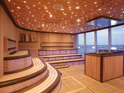 Moderne Holzsauna auf der Carnival Luminosa. Geschwungene Bänke mit indirekter Beleuchtung, Sternenhimmel-Decke und große Fenster mit Meerblick. Rechts ein Saunaofen.