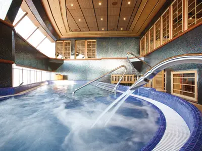 Geräumiger Innenpool mit blauen Mosaikfliesen, sprudelndem Wasser und mehreren Wasserstrahlen. Metallische Handläufe führen ins Becken. Seitliche Fenster, eine Holzbank und dezente Beleuchtung runden das Bild ab.