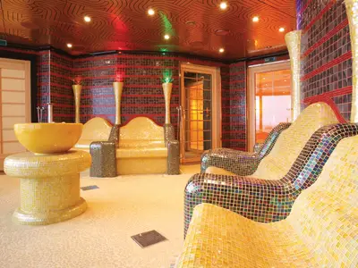 Ein luxuriöser Spa-Raum auf der Carnival Luminosa. Mehrere bequeme, mosaik-geflieste Liegen in Gelb- und Brauntönen. Ein gelbes Waschbecken auf einem Podest. Die Wände sind mit roten und braunen Mosaikfliesen bedeckt. Rotes und grünes Stimmungslicht an der Decke.