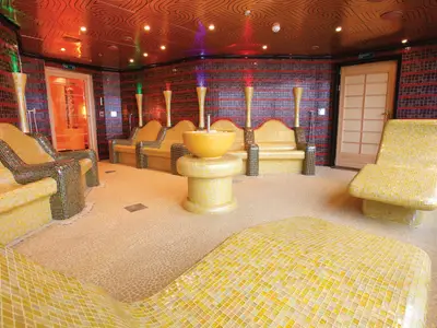 Ein Innenraum auf der Carnival Luminosa, gestaltet als Spa-Ruheraum. Mehrere gelb-schwarz geflieste Liegen und Stühle stehen um einen zentralen Eisbrunnen. Die Wände sind dunkelrot und schwarz gefliest, die Decke ist gemustert mit farbigen Lichtern. Eine Tür ist im Hintergrund zu sehen.