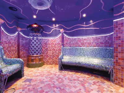Ein luxuriöser Spa-Raum mit lila-blauem Ambiente. Mosaikfliesen bedecken Wände, Böden und geschwungene Liegen. Ein zentraler Ofen und LED-Lichter schaffen eine entspannende Atmosphäre.