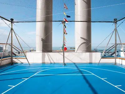 Ein blauer Multifunktions-Sportplatz mit weißen Linien auf dem oberen Deck der Carnival Luminosa, umgeben von hohen Schutznetzen. Im Hintergrund sind große weiße Schornsteine und das Meer mit einem weiteren Schiff zu sehen. Diverse Signalflaggen hängen an einem Mast.