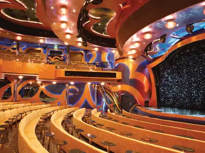 Weitwinkelansicht eines leeren Theaters auf der Carnival Luminosa. Reihen von beigen Sitzbänken mit Tischen blicken auf eine beleuchtete Bühne mit funkelndem Sternenhintergrund. Die Wände sind in warmen Orange- und Blautönen gehalten, mit vielen Lichtern und einer Treppe.