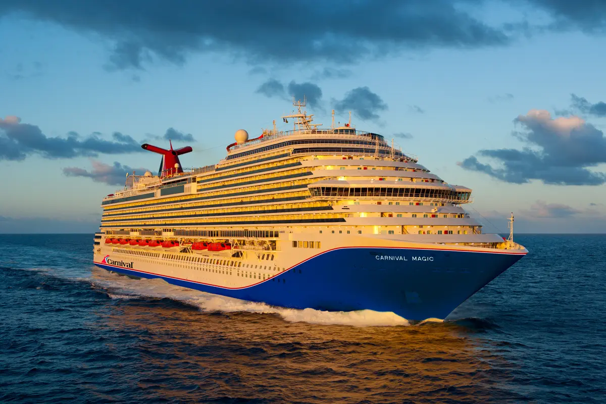 Die Carnival Magic auf dem offenen Meer, von der Seite aufgenommen. Der blaue Bug und das rote Carnival-Logo sind gut sichtbar. Die Sonne beleuchtet die Balkone. Leichte Wolken am Himmel.
