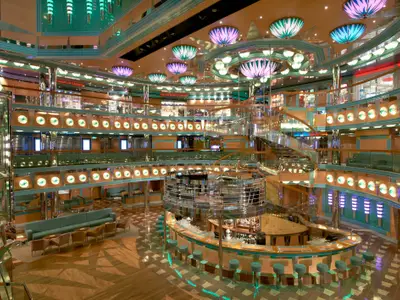 Blick von oben auf das mehrstöckige Atrium der Carnival Magic. Eine geschwungene Glas- und Chromtreppe führt hinunter zu einer großen, runden Bar. Ringsum sind Balkone mit Lounge-Bereichen und Geschäften zu sehen. Zahlreiche bunte Decken- und Wandleuchten illuminieren den Raum.