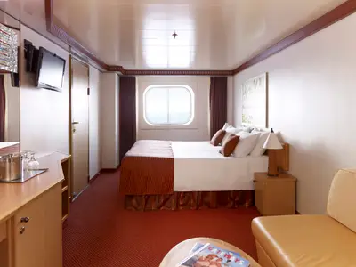 Innenansicht einer geräumigen Außenkabine auf der Carnival Magic mit Doppelbett, runder Fensteröffnung und Blick aufs Meer. Warme Brauntöne und rote Teppichböden prägen das Interieur.