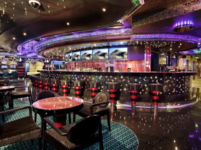 Moderne Bar auf der Carnival Magic. Leuchtende Theke, rote Barhocker und Bildschirme mit Schiffsansichten. Im Hintergrund Spielautomaten.