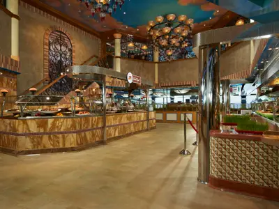 Geräumiges Buffet-Restaurant an Bord der Carnival Magic. Es gibt mehrere Essensstationen mit vielseitiger Auswahl, eine große Treppe und eine markante Deckenbeleuchtung. Der Raum ist warm und einladend gestaltet.