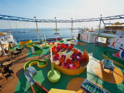 Bunter Sport- und Aktivitätsbereich auf dem Oberdeck der Carnival Magic bei Tageslicht. Zu sehen sind Minigolfbahnen, ein Seilgarten, zahlreiche farbige Stühle in einem Lounge-Bereich, ein Bildschirm mit dem "Sport Square"-Logo, Tischfußballtische und Tische mit Stühlen.