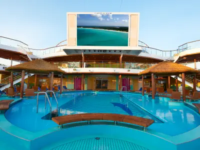 Weitwinkelansicht des Pools an Bord der Carnival Magic. Der Poolbereich umfasst mehrere Ebenen mit Strohdach-Bars und einem großen LED-Bildschirm, der eine karibische Bucht zeigt. Um den Pool herum gibt es Sitzgelegenheiten und Liegestühle. Die oberen Decks sind über Treppen erreichbar.
