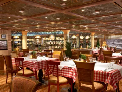 Ein festlich gedecktes Restaurant auf der Carnival Magic mit vielen Tischen, rot-weiß karierten Tischdecken und hölzernen Stühlen. Die Decke ist mit dekorativen Kupferplatten verziert. Im Hintergrund sind dunkle Weinflaschen und schmiedeeiserne Dekorationen zu sehen.
