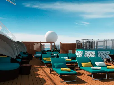 Blick auf ein sonniges Schiffsdeck mit mehreren Reihen von türkisfarbenen Liegestühlen und runden Himmelbetten. Im Hintergrund sind das Meer und ein großes weißes Radom zu sehen. Der Himmel ist blau.