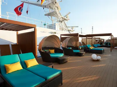 Sonniges Deck der Carnival Magic mit modernen türkisfarbenen Lounge-Möbeln und Liegen auf Holzdeck. Pergolen und weißer Aufbau erkennbar.