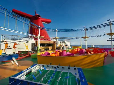 Ein lebhaftes Sport- und Unterhaltungsdeck auf der Carnival Magic. Im Vordergrund sind zwei Kickertische und eine Minigolfbahn zu sehen. Ein farbenfroher Sitzbereich mit vielen Stühlen umgibt eine tiefe gelbe Grube. Im Hintergrund erstreckt sich ein Hochseilgarten unterhalb eines großen roten Schornsteins. Eine Bar und das weite Meer sind ebenfalls sichtbar, alles unter einem klaren blauen Himmel.