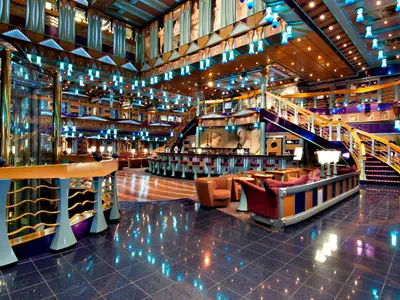 Eine lebhafte, mehrstöckige Bar-Lounge der Carnival Miracle. Der Raum ist in hellen blauen und orangefarbenen Tönen dekoriert und bietet eine geschwungene Bar mit Hockern, verschiedene Sitzbereiche mit Sesseln und eine große Treppe auf der rechten Seite. Blaue Fliesen reflektieren die Beleuchtung.