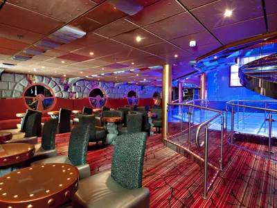 Innenansicht einer modernen Lounge der Carnival Miracle. Dunkle Sessel und runde Tische auf rotem Teppich. Links Steinwand, rechts blaue Wendeltreppe.