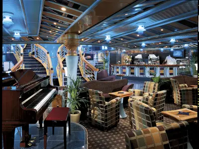 Blick in eine stilvolle Bar-Lounge der Carnival Miracle mit Grand Piano, dekorativer Treppe, verschiedenen Sitzbereichen und einer Bar mit blauer Deckenbeleuchtung.