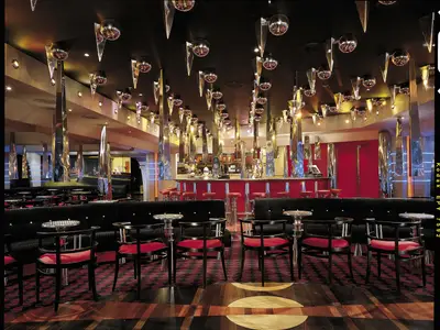 Innenansicht einer opulenten Bar-Lounge. Ein langer, roter Bartresen mit Hockern erstreckt sich im Hintergrund. Dunkle Sitzgruppen mit roten Stühlen stehen im Vordergrund. Die Decke ist mit vielen spiegelnden Kugeln und länglichen, glänzenden Skulpturen dekoriert, die das Licht reflektieren. Ein roter Teppich mit Muster bedeckt den Boden. Keine Personen sichtbar.
