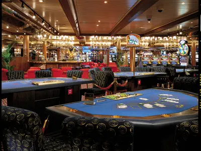 Blick in ein elegantes Casino auf der Carnival Miracle mit zahlreichen blau befilzten Spieltischen, darunter Blackjack- und Pokertische, sowie Reihen von Slot-Maschinen im Hintergrund. Goldene Kronleuchter und indirekte Beleuchtung sorgen für eine luxuriöse Atmosphäre. Die Stühle an den Tischen sind rot oder mit einem gold-schwarzen Spiralmuster bezogen.