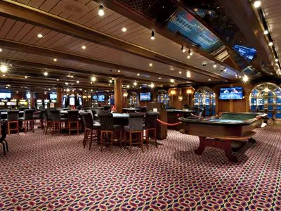 Elegantes Casino auf der Carnival Miracle. Sichtbar sind Spieltische wie Roulette mit Barhockern und Reihen von Spielautomaten. Der Raum ist luxuriös gestaltet mit Holzvertäfelungen, gemustertem Teppich und vielen hellen Spotlights.