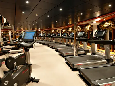 Ein geräumiger, gut ausgestatteter Fitnessraum auf der Carnival Miracle mit modernen Life Fitness Laufbändern und Fahrrädern. Der Raum hat dunkle Decken, reflektierende Oberflächen, indirekte Beleuchtung und große Fenster mit goldenen Vorhängen, die nach außen blicken.