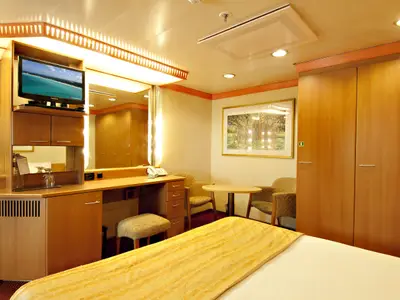 Helle, moderne Kabine der Carnival Miracle. Helles Holzinterieur mit Schminktisch, TV, Kleiderschrank, kleiner Sitzecke mit Tisch und Stühlen. Bett im Vordergrund.