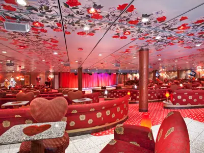 Opulent gestaltete Theater-Lounge auf der Carnival Miracle im Alice im Wunderland Stil. Der weite Innenraum ist rot dominiert mit herzförmigen Stühlen und rosengemusterten Sofas. Eine Bühne mit roten Vorhängen ist im Hintergrund sichtbar. Die Decke zeigt rote Rosen und indirekte Beleuchtung. Überall sind Themendekorationen und viele Sitzgelegenheiten.