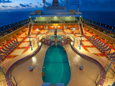 Blick von oben auf das beleuchtete Pooldeck der Carnival Miracle bei Abenddämmerung. Ein großer Pool, ein Whirlpool mit Skulptur und viele Liegestühle sind zu sehen. Lichter auf dem Wasser in der Ferne.