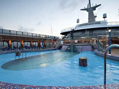 Abendliche Ansicht des großen blauen Pools auf dem Oberdeck der Carnival Miracle. Liegestühle, Balustraden und der Schiffsaufbau mit Radarantennen und einer Statue sind sichtbar.