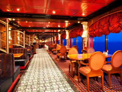 Ein luxuriöses Restaurant auf der Carnival Miracle mit langen Reihen von Sitzkabinen und Tischen am Fenster, die einen weiten Blick auf das blaue Meer bieten. Rote Decken und Vorhänge prägen die Einrichtung.