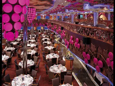 Blick in ein zweistoeckiges Restaurant der Carnival Miracle mit vielen Tischen und Stühlen, eingedeckt für Gäste. Pinke, kugelfoermige Dekorationen zieren Wände und Säulen. Ein oberer Balkon mit weiteren Sitzplaetzen ist sichtbar.