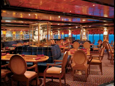 Großzügiges, elegantes Schiffsrestaurant mit dunklen Samt-Sitzbänken und Holztischen. Im Zentrum steht ein Modell eines Segelschiffs in einer Glasvitrine. Große Fensterfront bietet Blick auf das blaue Meer.