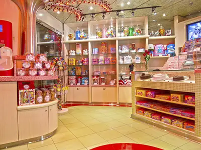 Ein heller Süßwarenladen auf der Carnival Miracle. Regale mit bunten Bonbons, Lollipops und Schokolade, sowie ein Display mit Torten, sind sichtbar.