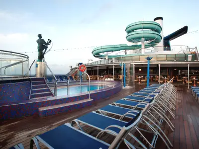 Blick auf das leere Sonnendeck der Carnival Miracle mit Pool, blauen Liegestühlen, einer Wasserrutsche und einer Skulptur. Im Hintergrund eine Bar und das offene Meer.