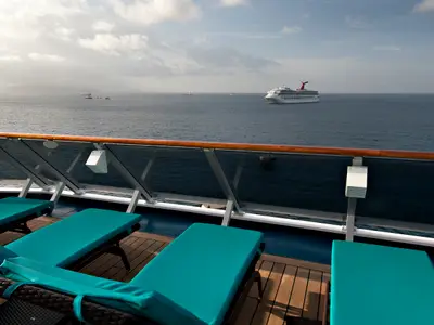 Eine Ansicht von einem Sonnendeck der Carnival Miracle, mit mehreren türkisfarbenen Liegestühlen im Vordergrund. Dahinter erstreckt sich das weite Meer mit einem weiteren großen Schwestersschiff und kleineren Schiffen am Horizont unter einem bewölkten Himmel.