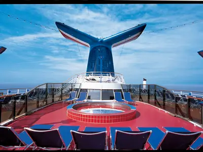 Das Sonnendeck der Carnival Miracle mit einem runden Whirlpool und vielen blauen Liegestühlen unter dem markanten Heckschornstein. Blauer Himmel und Meer im Hintergrund.