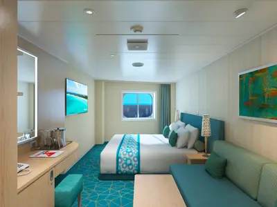 Helles, modernes Kabinen-Interieur der Carnival Panorama mit Doppelbett, türkisfarbenem Sofa, Schreibtisch und einem runden Fenster mit Blick auf das blaue Meer und einen bewölkten Himmel.