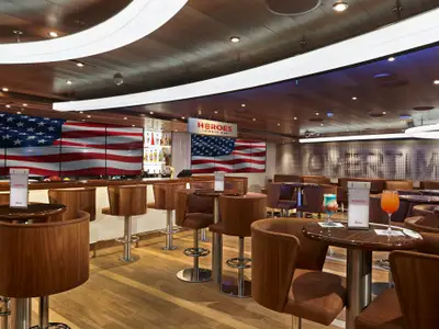Innenansicht der 'Heroes Tribute Bar' auf der Carnival Panorama mit einer großen amerikanischen Flagge auf Videobildschirmen, Holzeinrichtung, bequemen Stühlen und Cocktails auf den Tischen.