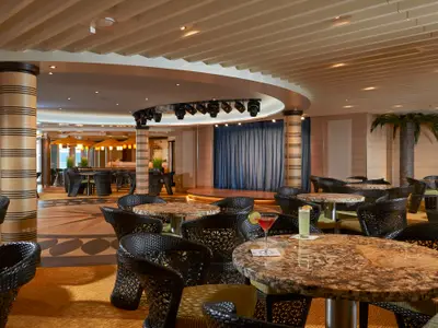 Innenansicht einer stilvollen Bar-Lounge auf der Carnival Panorama. Dunkle Rattanstühle, Granit-Tische, eine Bühne und eine Palmen-Dekoration schaffen eine entspannte Atmosphäre.