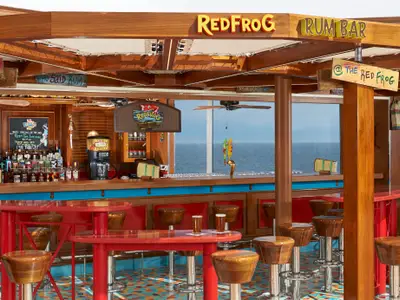Ein farbenfrohes, karibisch dekoriertes Bar-Interieur mit Holztresen, roten Tischen und Hockern. Große Fenster bieten einen weiten Blick auf das blaue Meer. Die Bar ist leer, mit Flaschen und Zapfanlagen bereit.