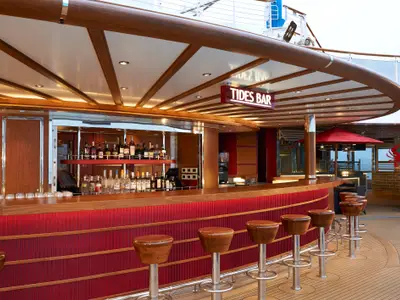 Eine moderne Outdoor-Bar namens 'TIDES BAR' auf der Carnival Panorama mit rotem Tresen, Holzstühlen und Blick auf das Meer. Überdachtes Deck mit Flaschenregalen und Sitzbereichen im Hintergrund.