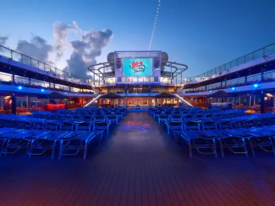 Außendeck der Carnival Panorama bei blauer Stunde. Blaue Liegestühle auf Holzdeck vor großem LED-Bildschirm mit 'Dive-In Movies' Logo. Blaue Lichter schmücken die Decks. Ohne Personen.