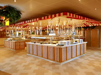 Ein hell beleuchtetes Buffetrestaurant an Bord der Carnival Panorama mit mehreren Anrichteinseln voller Speisen und Tellern unter rot-weiß gestreiften Markisen. Kuenstliche Palmen schmücken den Raum.