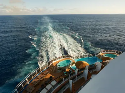 Heckansicht der Carnival Panorama: Meer mit Kielwasser. Sonnendeck mit Pools, Liegen und strohgedeckten Schirmen. Blauer Himmel, leichte Bewölkung.