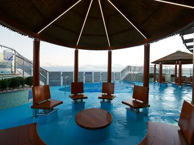 Ein überdachter Poolbereich auf der Carnival Panorama mit Holzsitzen und Tisch im Wasser. Dahinter weitere Pools, Sonnendecks und das offene Meer.