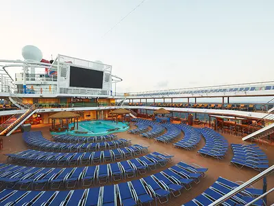 Sonnendeck der Carnival Panorama mit Pool, blauen Liegestühlen und großem Bildschirm. Mehrere Decks im Hintergrund. Keine Personen.