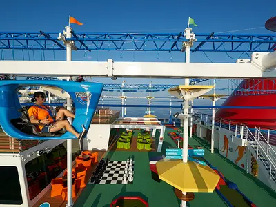 Eine Person sitzt in einem blauen SkyRide-Gondelwagen hoch über dem Deck der Carnival Panorama, während im Hintergrund ein Hochseilgarten, eine Minigolfanlage und ein riesiges Schachspiel sichtbar sind, mit weitem Blick auf das Meer unter blauem Himmel.
