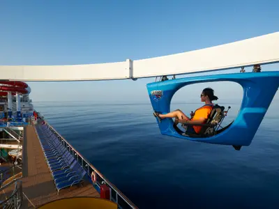 Eine Person fährt auf einer blauen SkyRide-Gondel, die über das Meer hinausragt. Im Hintergrund ist das Sonnendeck der Carnival Panorama mit roten Wasserrutschen und vielen blauen Liegestühlen zu sehen. Blauer Himmel und ruhiges Meer bilden die Kulisse.