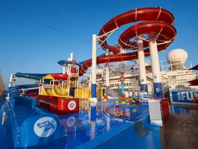 Detailansicht des Carnival WaterWorks Wasserparks an Bord der Carnival Panorama. Mehrere rote und blaue Wasserrutschen schlängeln sich über ein farbenfrohes Planschbecken mit interaktiven Wasserspielen unter strahlend blauem Himmel. Eine große weiße Radarkuppel des Schiffs ist im Hintergrund sichtbar.